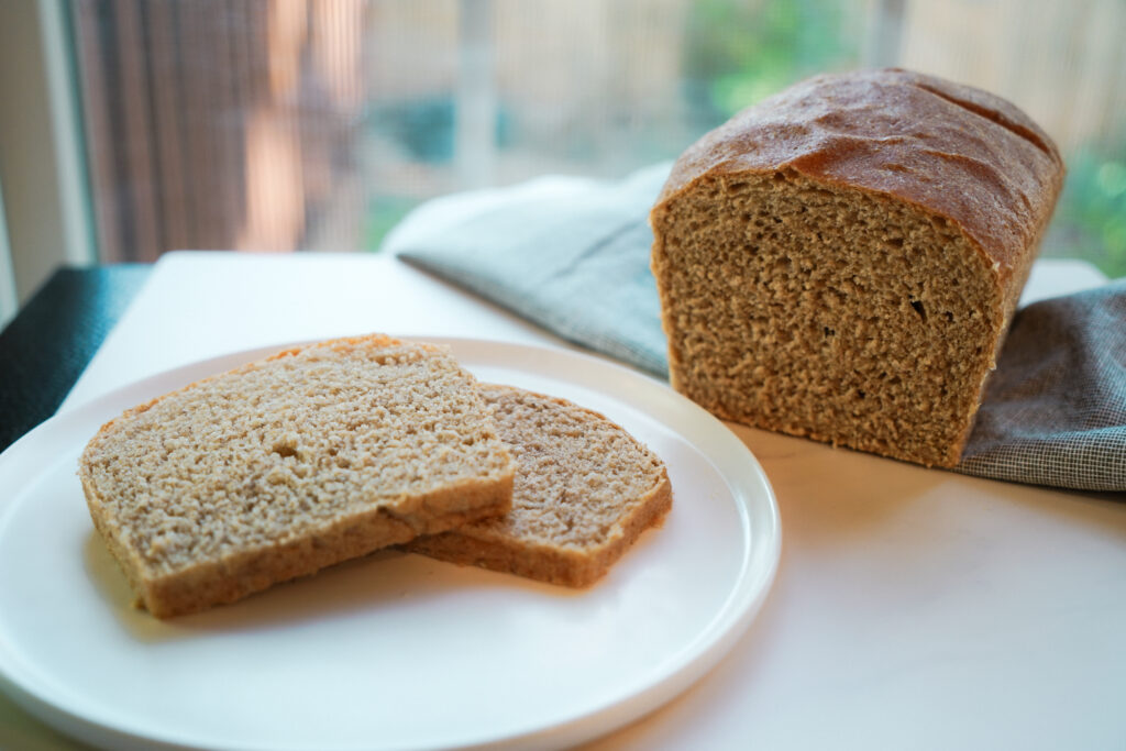 Two slices of freshly milled honey wheat bread and a cut loaf of bread on a white table